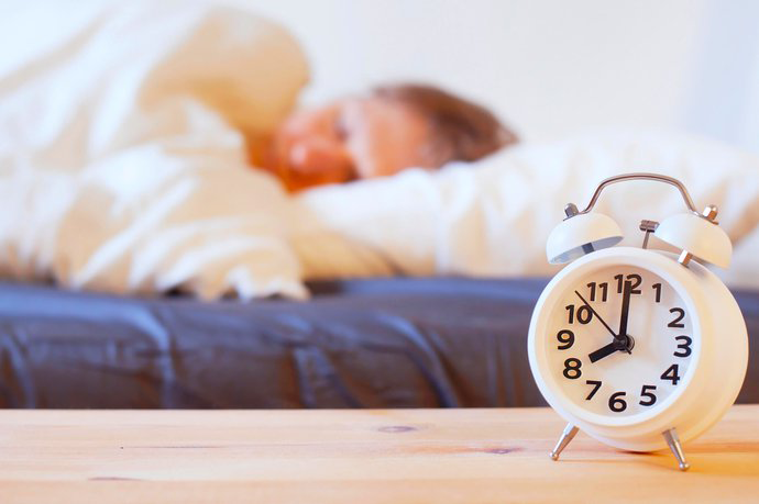 White alarm clock with black hands and numbers to right foreground, blurred woman sleeping behind on blow-up bed